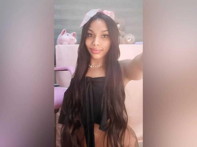 Amaranta69s with long black hair, wearing a black top, necklace, and pink bow, showing her bare ass and pouty lips, standing between pink and gray walls.