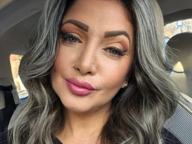 Miky69I has gray wavy hair, pink lipstick, and thick eyebrows with gold eyeshadow and long lashes, sitting in a car with a subtle smile. Boobs not visible.