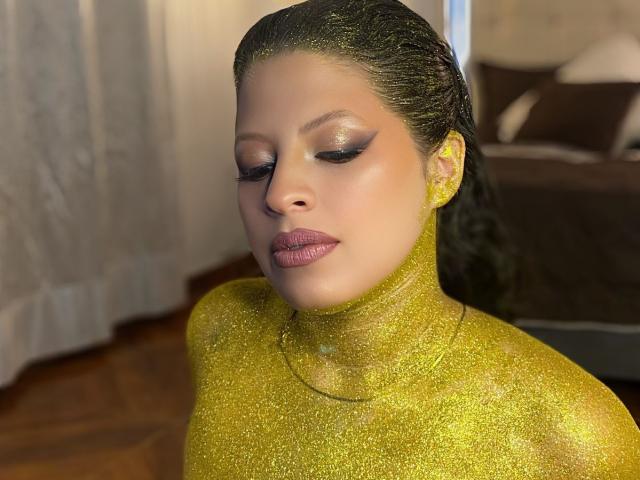ShyVibes with glittery yellow skin, closed eyes, and pink lipstick, has shiny dark hair slicked back. Boobs and face are covered in glitter, looking down with a calm expression.