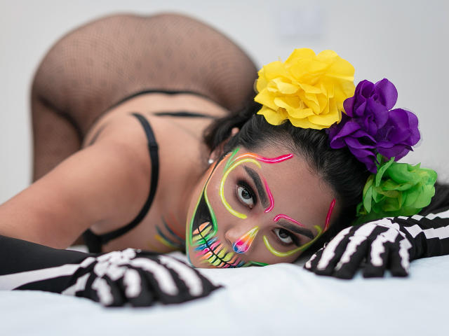 PaolaYKatty au lit, le visage maquillé avec un motif coloré de Día de los Muertos, des fleurs jaunes, violettes et vertes dans les cheveux, des bas en maille noire, un soutien-gorge noir, des gants squelette noirs, fesses relevées..