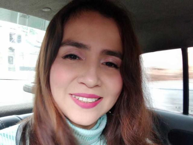 HeidyWells has pink lipstick, brown eyes, and long brown hair, smiling in a car with a light blue turtleneck, showing her cute ass (tits) in the background.