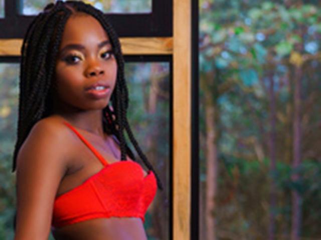 LewaChasco, con la pelle scura, indossa un reggiseno di pizzo rosso, è in piedi vicino a una finestra, guarda di lato con i capelli intrecciati, mostrando il seno e un'espressione sottile e sensuale..