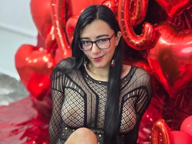 JewelryQueen with long black hair, glasses, and red lipstick, wearing a see-through black mesh top, exposing her tits. Red heart balloons in the background.