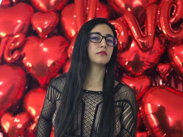 JewelryQueen mit langen, schwarzen Haaren, die eine Brille trägt und ein durchsichtiges schwarzes Kleid anhat, steht vor roten, herzförmigen Ballons und glänzenden "LIEBE"-Lettern und wirkt leicht gelangweilt..