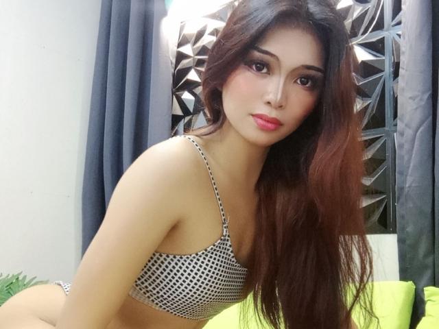 QueenMetina with long brown hair, wearing a black and white polka dot bra, leaning forward, showing her tits, pink lipstick, and dark eye makeup, in a bedroom setting.