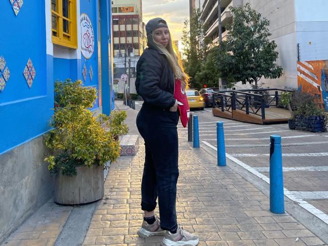 JennyLawry è in piedi su un marciapiede cittadino, indossa una giacca nera, un berretto e pantaloni neri, tiene in mano una maglietta rossa e porta delle scarpe con il tacco a zeppa, guardandosi indietro per le spalle..