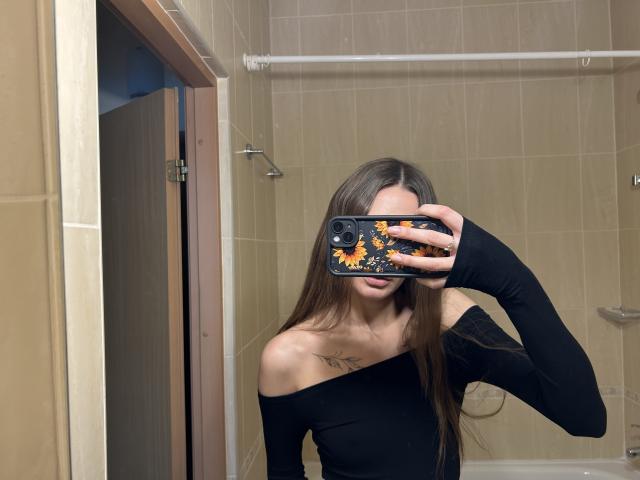 Karish with long brown hair, black off-shoulder top, holding phone with sunflower case, covering face, small tattoo on chest, in bathroom mirror selfie, showing her tits.