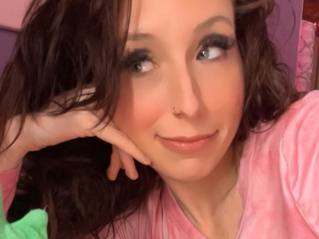 TinaLocket has wavy brown hair, dark eyeliner, a nose ring, and a pink shirt, smiling slightly with her hand on her cheek, looking to the side.