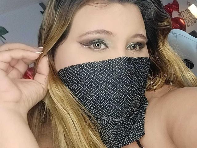 SalmaAzaman with blonde-to-brown ombre hair, green eye makeup, and black patterned mask covering her nose and mouth, holding her hair with a white-painted nail.