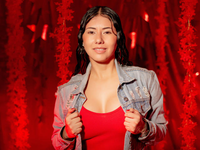 XiaQueen mit nassen, schwarzen Haaren, trägt ein rotes Oberteil und eine blaue Jeansjacke, hält die Ränder der Jacke fest, lächelt, zeigt kleine Brüste, vor einem roten, blättrigen Hintergrund..