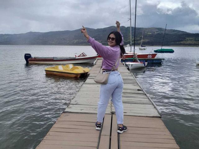 FrancheskaWaalsh stands on a wooden dock, arms raised, wearing a pink shirt, white pants, black shoes, and sunglasses. Boats are anchored in the calm water.