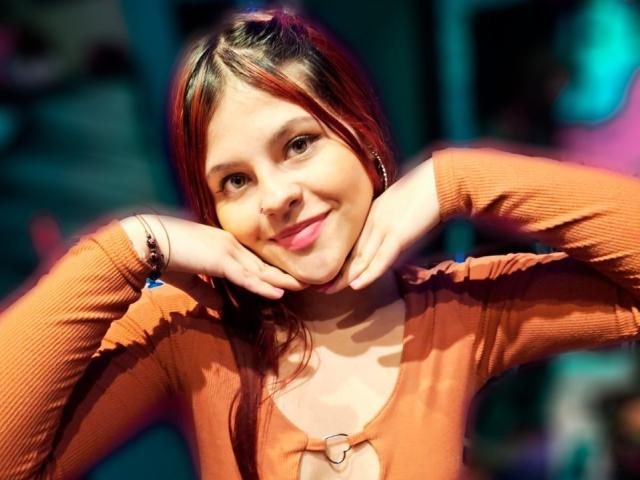 SophieHill with red hair, smiling, hands on cheeks, wearing an orange ribbed top with a heart-shaped cutout, bracelet on right wrist, soft lighting, slight bokeh effect.