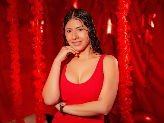 XiaQueen mit feuchten, braunen Haaren, trägt ein rotes Tanktop, zeigt ihre Brüste, lächelt, Hand am Kinn, roter Federhintergrund, rote Armbanduhr, sieht sexy aus, helle Haut, süß..