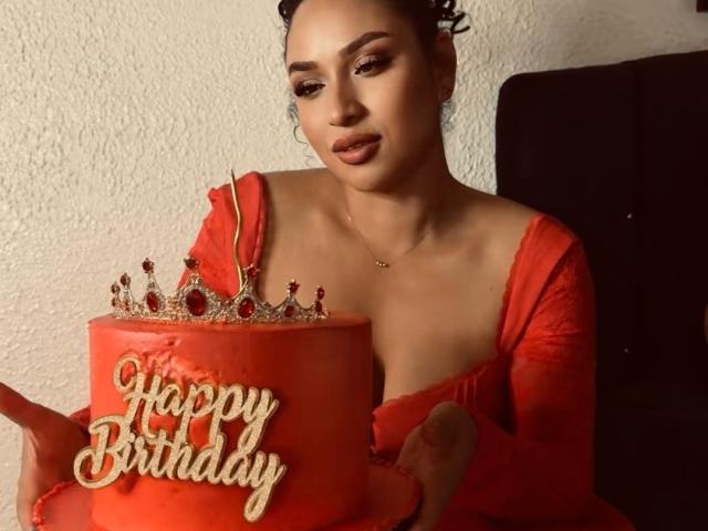 CarlaMuille with curly hair, wearing a red off-shoulder dress, holding a red cake with a jeweled crown and "Happy Birthday" in cursive. Her tits are slightly visible.