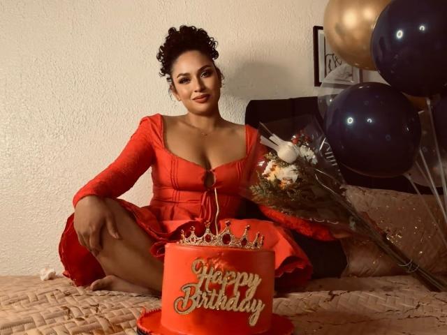 CarlaMuille with curly black hair, red dress, and large tits, sitting on a bed with a red birthday cake, bouquet, and balloons. She's smiling, with her legs spread.