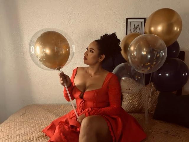 CarlaMuille with dark curly hair in a red dress, holding a gold balloon, sitting on a bed with gold and clear balloons, her big tits visible, looking to the side.
