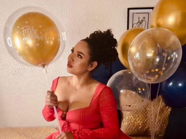 CarlaMuille, con i capelli ricci, indossa un vestito rosso, tiene in mano dei palloncini dorati, guarda di lato, con il seno leggermente visibile, seduta su una superficie beige, in una stanza accogliente..
