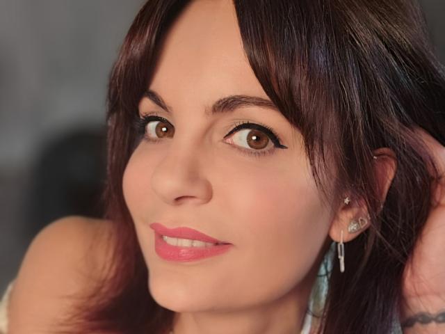 Close-up de JuneLaFrenchie, uma mulher de olhos castanhos, com cabelo castanho escuro, batom rosa e maquiagem discreta, olhando diretamente para... a câmera, um leve sorriso, usando brincos de diamante, uma luz suave realçando seu rosto.