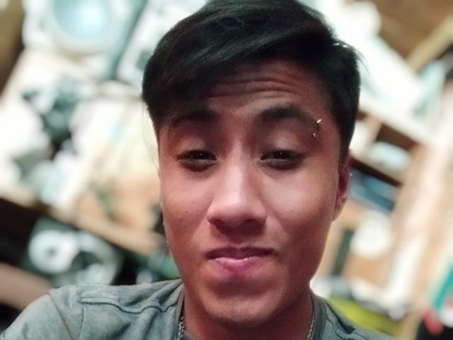AnthonySexyMan with short black hair, slight smile, small nose ring, and a grey shirt, looking into camera, slightly blurred background, medium skin tone, casual and relaxed.