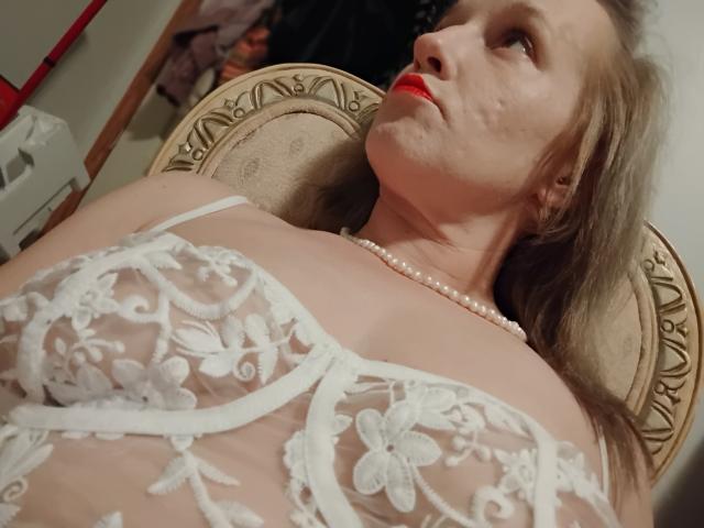Sortasassy with red lipstick, wearing white lace bra, pearl necklace, lying back in ornate chair, looking up with a slight smile, blonde hair, fair skin. Boobs visible through lace.