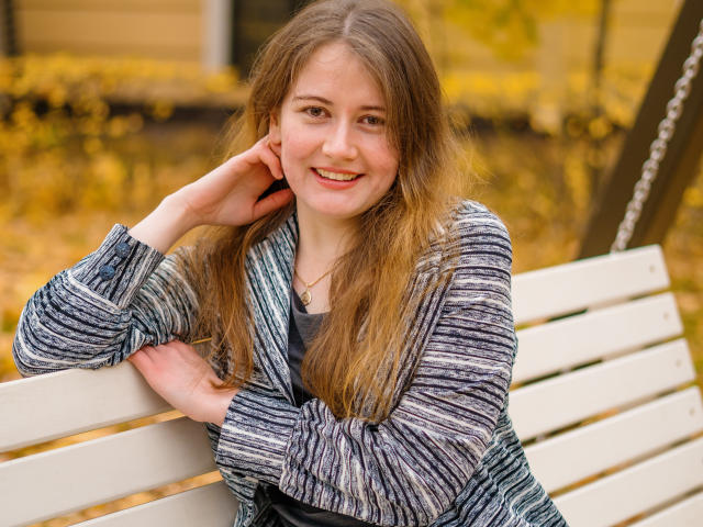 EvaNoNude with long brown hair, smiling, wearing a gray striped sweater, leaning on a white bench, right hand touching her cheek, autumn leaves in the background.