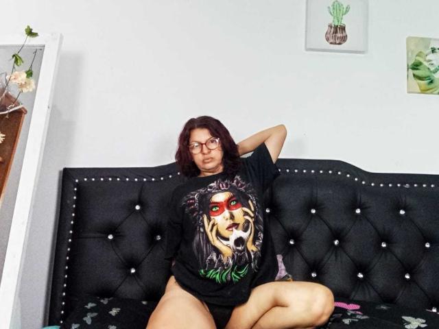 AntonellaDupontt, con cabello rojo y gafas, vestida con una camiseta negra con el logo de "Lioness", sentada en un sofá negro con capitonado, con las piernas separadas, mostrando su vulva. Su mano está apoyada en su cabeza..