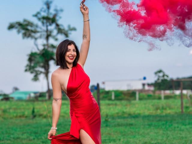 AnastasiaEvansX in a red one-shoulder dress, holding up a smoking piece of red cloth, smiling, with a tattoo on her arm, in a grassy field.