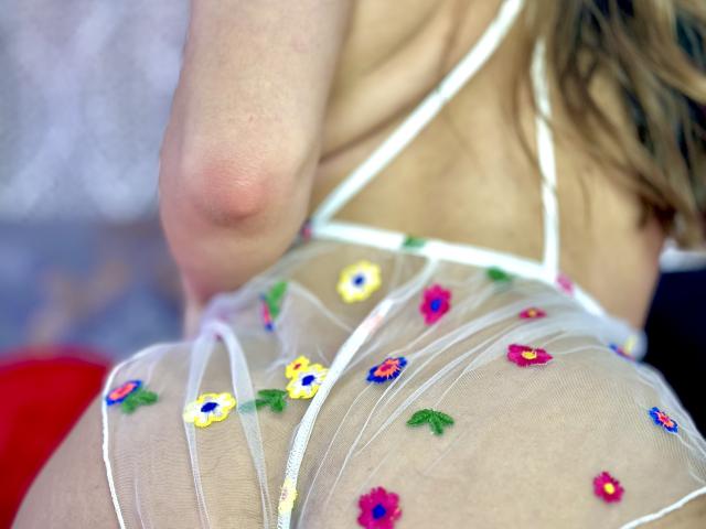 DouceMila's ass in a sheer white top with colorful embroidered flowers, showing her bare back and a partial view of her blonde hair. Her right arm is visible.