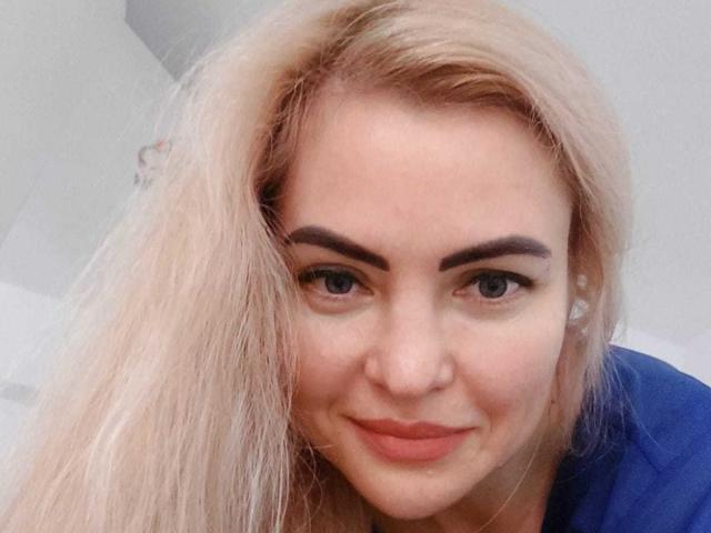 Laura69SexyHot with blonde hair, blue eyes, and a cute smile, wearing a blue shirt, has full eyebrows and slightly flushed cheeks, looking directly at the camera.