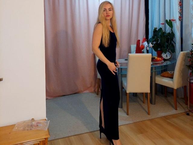 Laura69SexyHot, blonde, in black, tight, sleeveless dress with high slit, standing, holding dress, wood floor, white chairs, green plant, pink curtains, red vase, wooden table, heels.