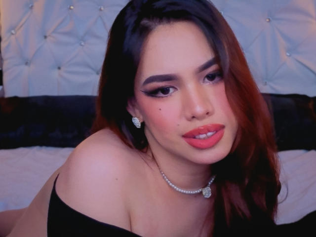 RogueCaliente with red hair, dark eyebrows, and pink lipstick, wearing a black off-shoulder top, diamond earrings, and necklace, lying on a bed, looking at the camera.