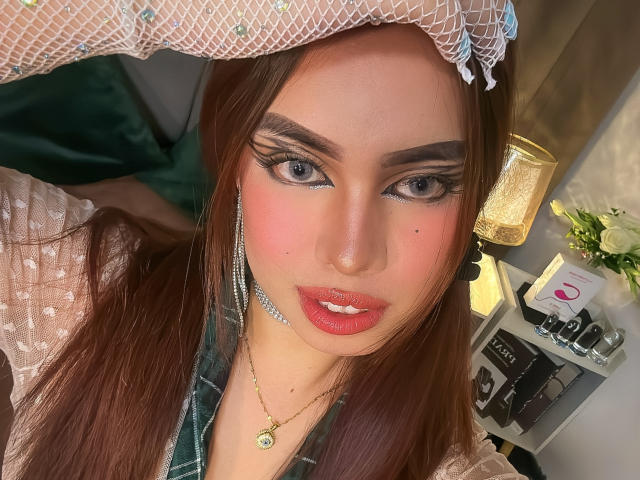 RogueCaliente has red lips, blue eyes, and a mole on her nose. She's wearing a lace headpiece, a green plaid shirt, and a gold necklace with a small green pendant.