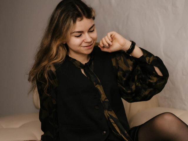 TinaRedly with wavy brown hair, wearing a black camo vest and striped shirt, sits on a beige couch, smiling softly, adjusting her wrist with a black bracelet.