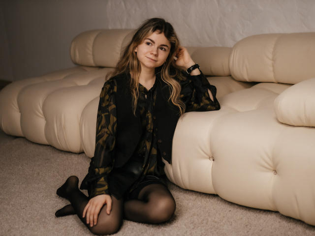 TinaRedly with wavy brown hair, sitting on beige carpet, wearing black sheer pantyhose, black dress, and patterned blouse, leaning on cream leather sofa, smiling slightly.