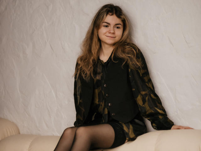 TinaRedly with wavy brown hair, wearing a black camo shirt, black vest, and black sheer stockings, sitting on a cream couch, smiling softly. Boobs not visible. Ass not visible.
