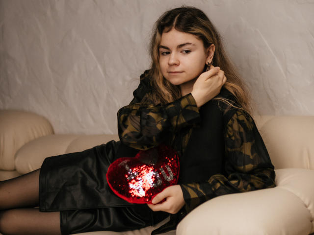 TinaRedly with wavy blonde hair, wearing a black dress and camo shirt, holds a red sequined heart, sitting on a beige couch, looking confident and slightly bored.