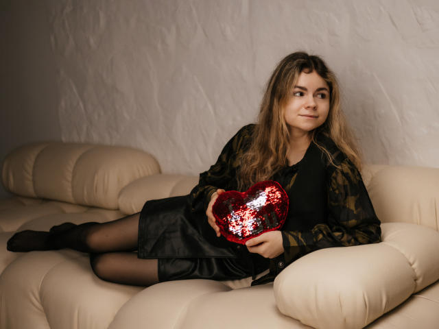 TinaRedly with long brown hair, black dress, and sheer black stockings, holding a red sequined heart, lounging on a beige leather couch. She's got a cute, relaxed smile.