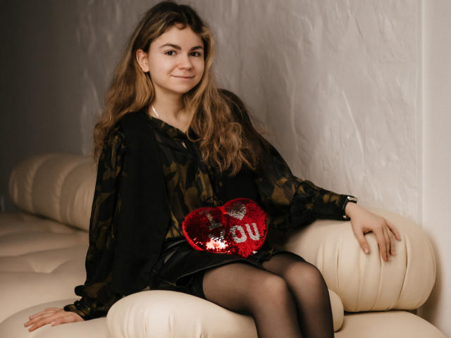 TinaRedly with long brown hair, wearing a black camo shirt, red sequined heart purse, black skirt, and black stockings, sitting on a cream leather couch.