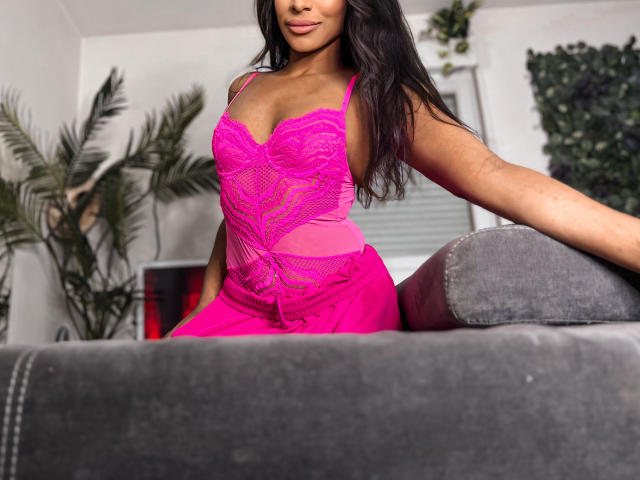 Ciarana with dark hair, pink lace and mesh lingerie, showing her tits and ass, sitting on a grey couch, arm resting on the back. Her skin is brown.
