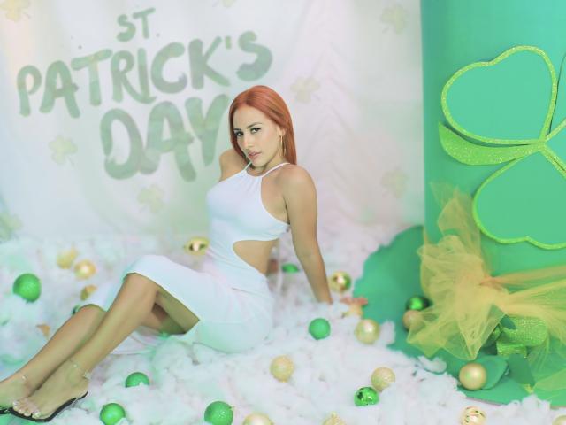 BombSophie with red hair, white cut-out dress, sitting on green and white ornaments, surrounded by fluffy white "grass," and a green shamrock. Her tits are small, her ass is round.