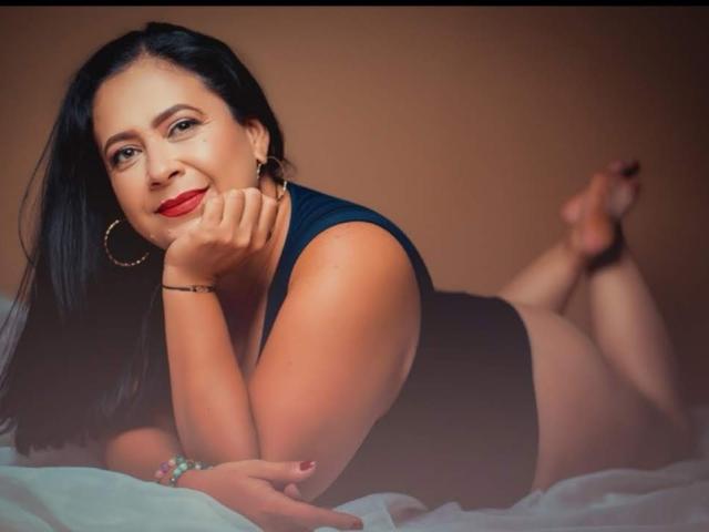 KarlaTom69 is a curvy woman with black hair, red lipstick, and hoop earrings, lying on a bed, wearing a black one-piece, her ass up, smiling at the camera.