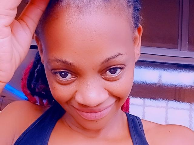HotMayaa with brown skin, small nose, and dark eyes, smiling slightly, holding her head with a hand, wearing a black halter top, and looking directly at the camera.