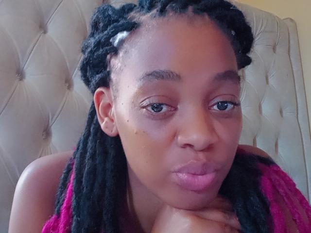 HotMayaa, avec sa peau brun foncé, ses cheveux noirs en tresses, ses lèvres roses et gonflées, et une expression sérieuse, repose son menton sur sa main. Elle porte un haut rose..