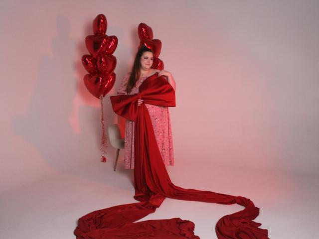 DaniellaGraceZZ with long brown hair, wearing a pink floral dress, holding a red satin blanket, standing beside red heart-shaped balloons, with a red blanket on the floor.