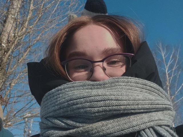 StaceyKiss with glasses, gray scarf wrapped around her face, black hoodie, and black-and-white hat, blue sky, bare trees in background.