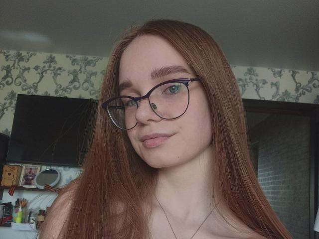 StaceyKiss with long red hair, glasses, and pale skin, showing off her tits in a selfie, wearing a thin necklace, smiling softly.