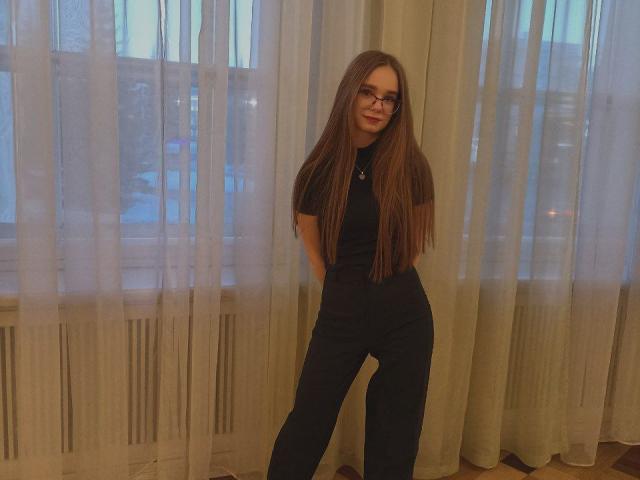 StaceyKiss with long brown hair, glasses, black t-shirt, and black pants, standing in front of sheer curtains, hands in pockets, smiling slightly, inside a softly lit room.