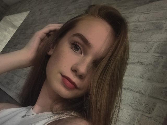StaceyKiss has fair skin, red lipstick, and long brown hair, with a subtle smile and hand in her hair. She's wearing a white top, looking at the camera.
