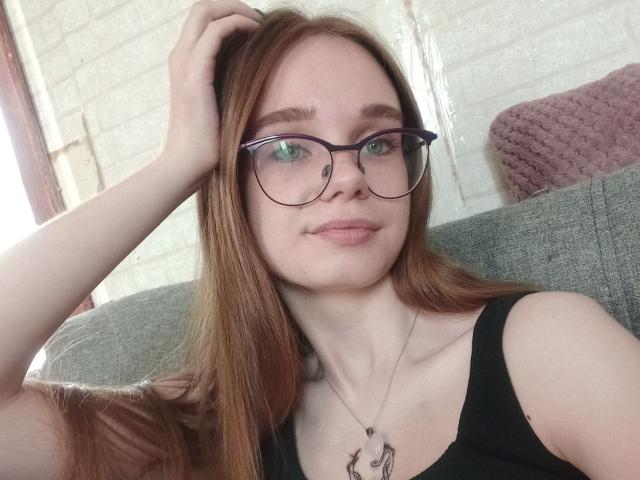 StaceyKiss with red hair, glasses, and black tank top, touching her head, showing her green eyes and subtle smile, wearing a necklace with a heart charm.