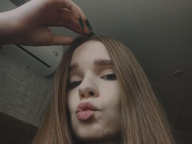 StaceyKiss has long brown hair, green nails, and is pouting her pink lips. Her hand is on her head. She has thick eyebrows and is looking down.